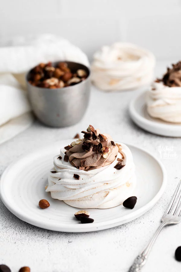 Individual Nutella Pavlovas on round white plates garnished with chopped hazelnuts and chocolate