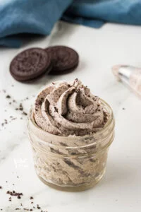 Cookies and Cream Frosting Recipe piped into a small glass jar with Oreo cookies and a blue kitchen towel in the background