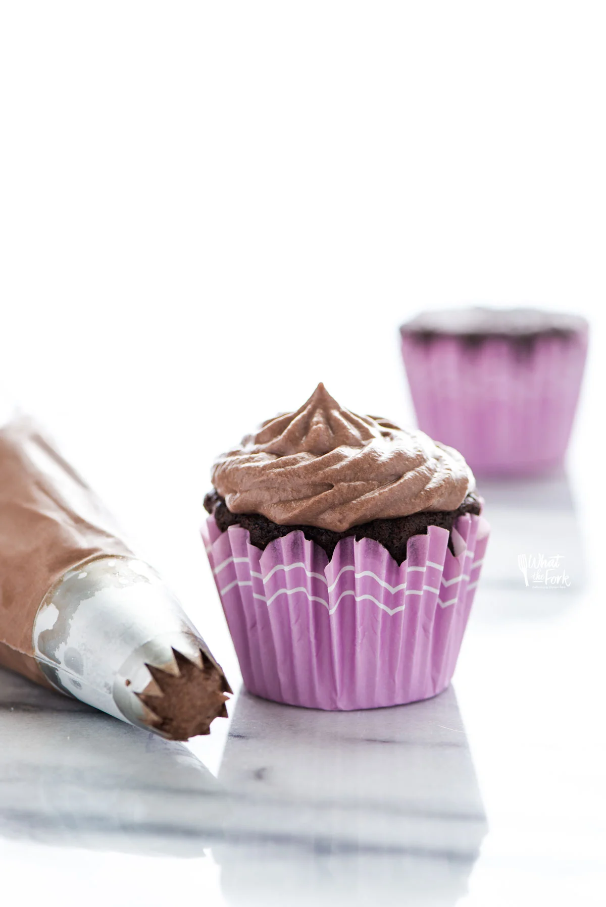 Nutella Whipped Cream piped onto a chocolate cupcake with a purple cupcake wrapper. The piping bag is fitted with a open star tip and next to the cupcake.