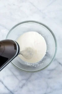 An electric hand mixer with a whisk attachment beating heavy cream in a large, clear glass bowl.