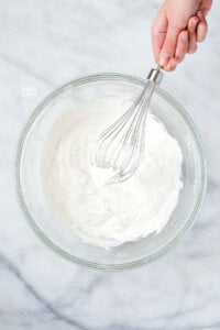 a hand holding a whisk with whipped cream holding stiff peaks over a bowl of whipped cream