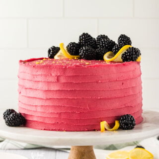 a gluten free lemon blackberry cake on a white a wood cake stand that is frosted with blackberry buttercream and decorated with fresh blackberries and lemon twists