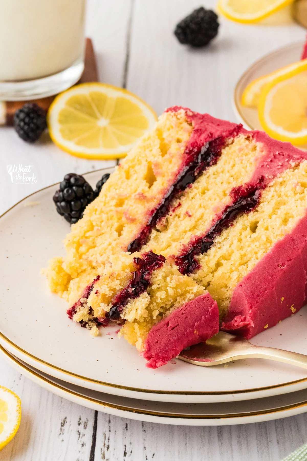 a slice of Gluten Free Lemon Blackberry Cake on a stack of round white plates with a forkful of cake on a gold fork
