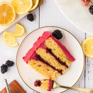 a slice of lemon cake with black berry frosting and blackberry cake filling on a round white plate. There is a fork of cake and the plate is surrounded by lemon slices and fresh blackberries.