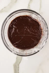 cooked chocolate pudding in a clear glass bowl with plastic wrap pressed directly onto the surface ready to be chilled