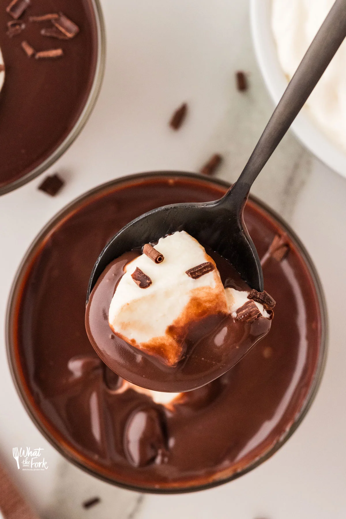 a spoonful of gluten free chocolate pudding with whipped cream and chocolate shavings held over a small clear glass cup filled with more chocolate pudding