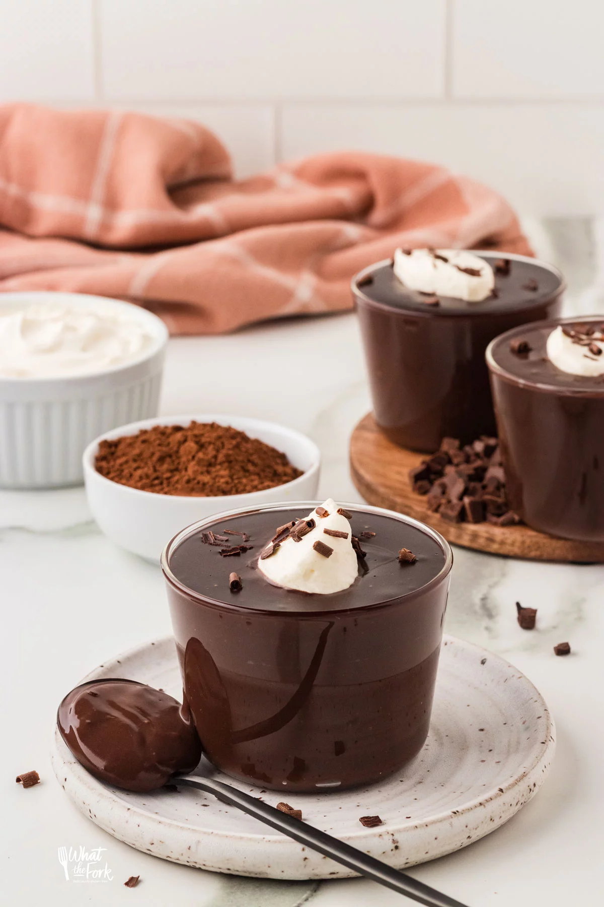 homemade chocolate pudding in a small clear glass cup topped with whipped cream and chocolate shavings. The cup is on a small round white pottery plate with a spoonful of chocolate pudding resting on the plate.