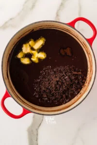 cooked gluten free chocolate pudding in a white and red Dutch oven. Cubed butter, chopped chocolate, and espresso powder have been added and are ready to be whisked in.