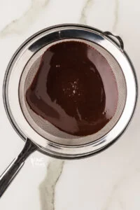 chocolate pudding being pressed through a fine mesh strainer over a glass bowl