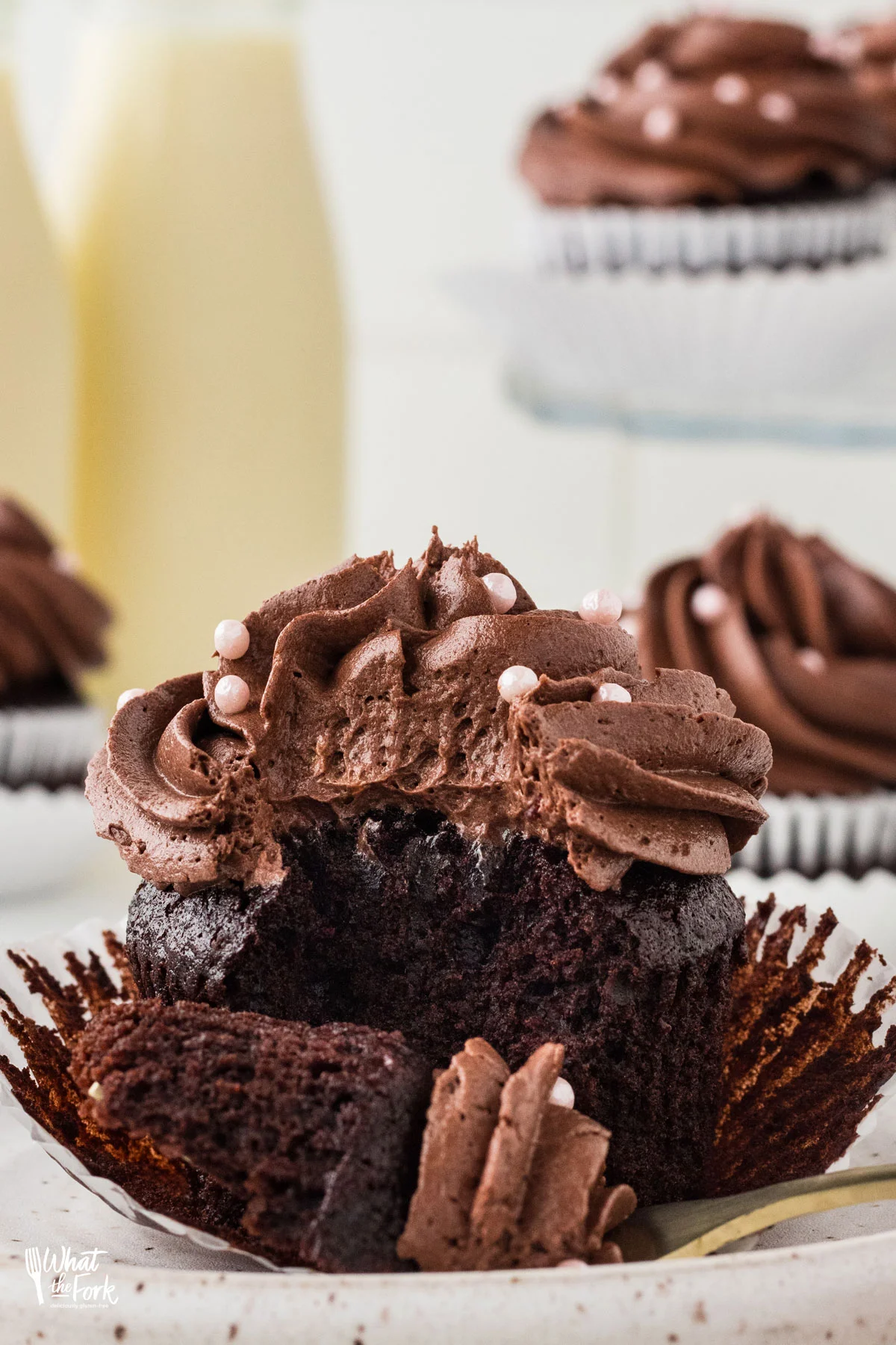 a moist gluten free chocolate cupcake with chocolate frosting on a white paper liner that has been opened. A gold fork is placed in front with a forkful of cupcake on it ready to be eaten.