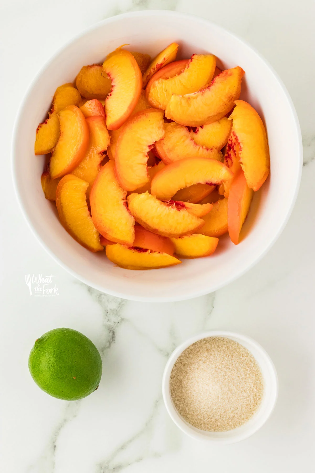Sliced peaches in a white bowl, sugar in a small white bowl, and a lime are set out to make macerated peaches.