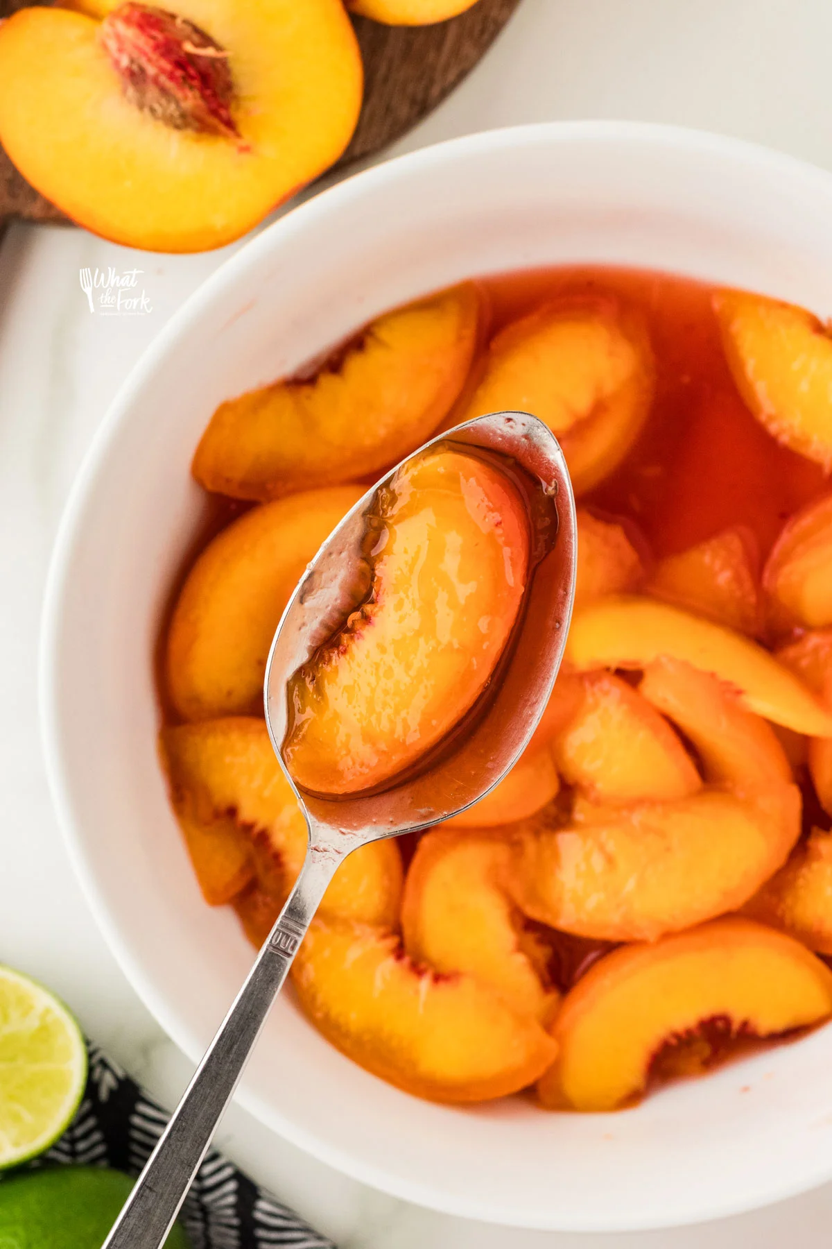 a silver spoon with peach slice and juice held over a white bowl full of macerated peaches