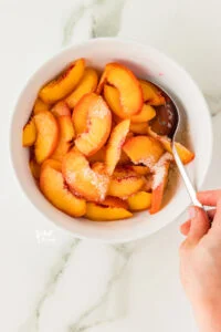 sliced peaches in a white bowl with granulated sugar and lime juice to make macerated peaches. A hand is holding a silver serving spoon to stir the peaches and sugar together.
