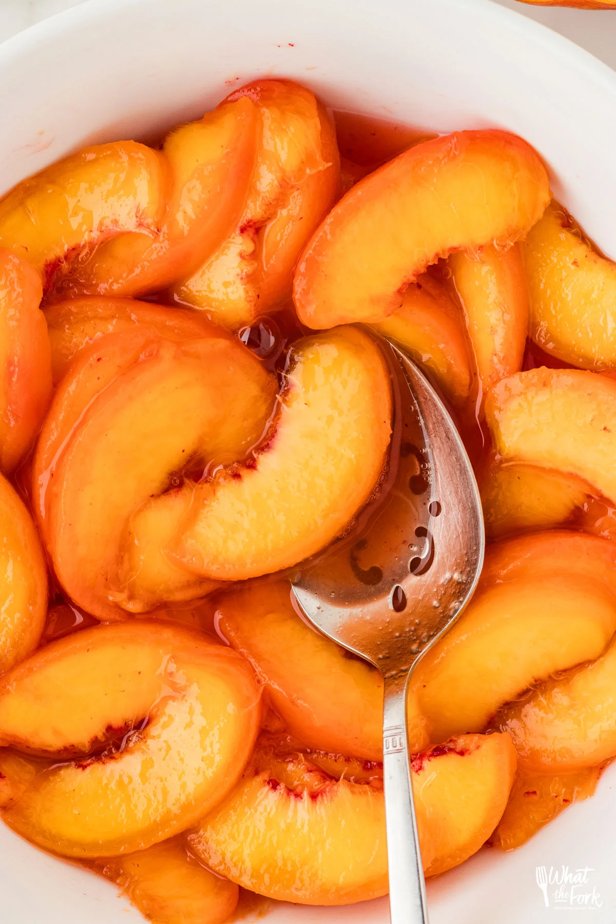 a white bowl of macerated peaches with a large silver serving spoon.