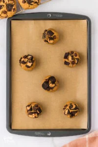 six balls of gluten free peanut butter chocolate chip cookie dough rolled and placed on a brown parchment paper lined cookie sheet