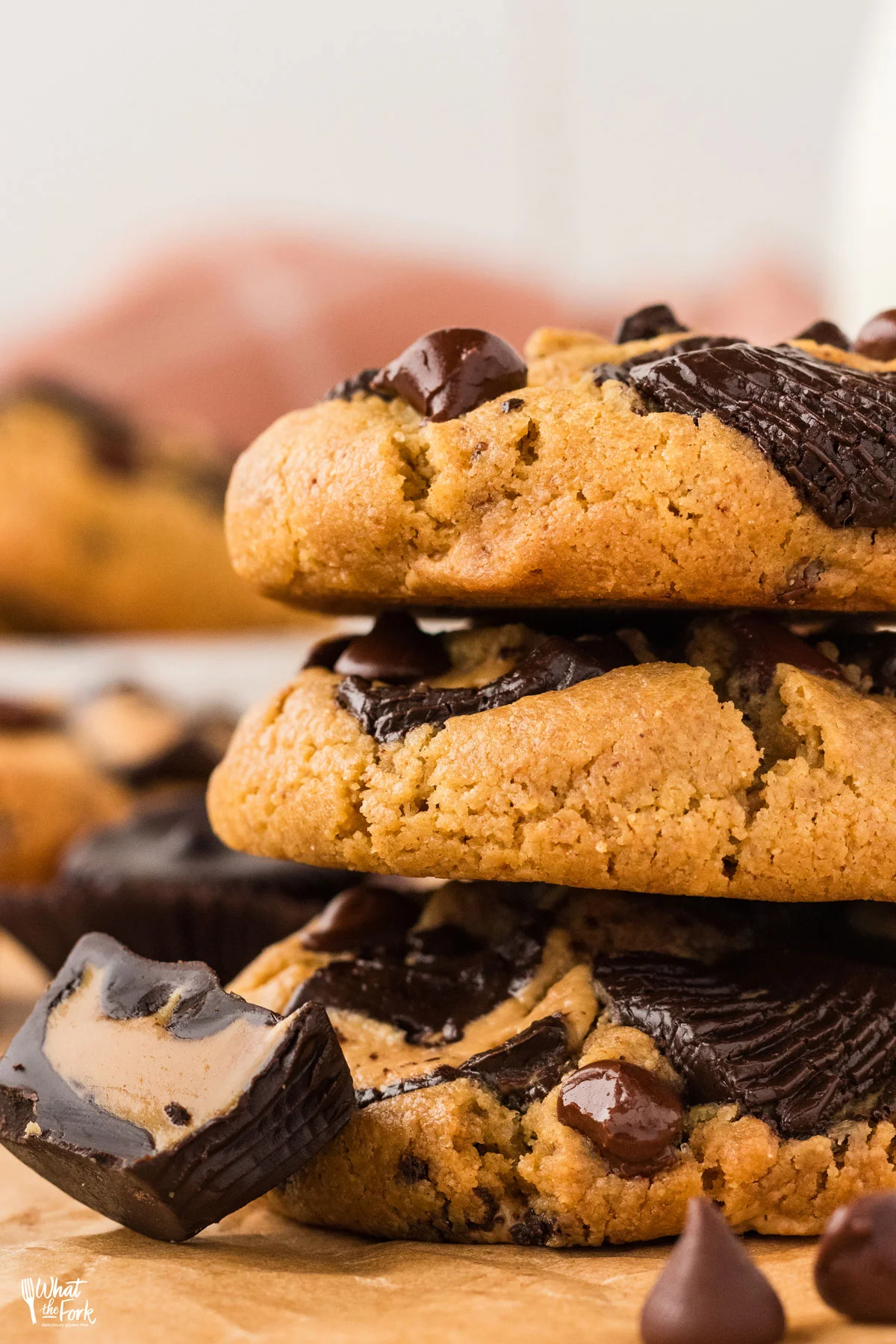 a stack of three baked Loaded Gluten Free Peanut Butter Chocolate Chip Cookies on brown parchment paper with half of a dark chocolate peanut butter cup leaning against it.