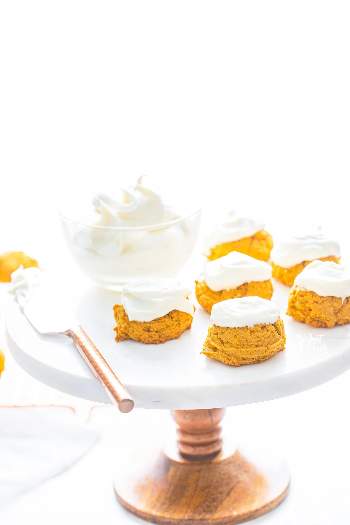 No Chill Gluten Free Pumpkin Cookies with Cream Cheese Frosting on a white cake with a wood base. The cake stand also has a small bowl of cream cheese frosting and knife to spread it.