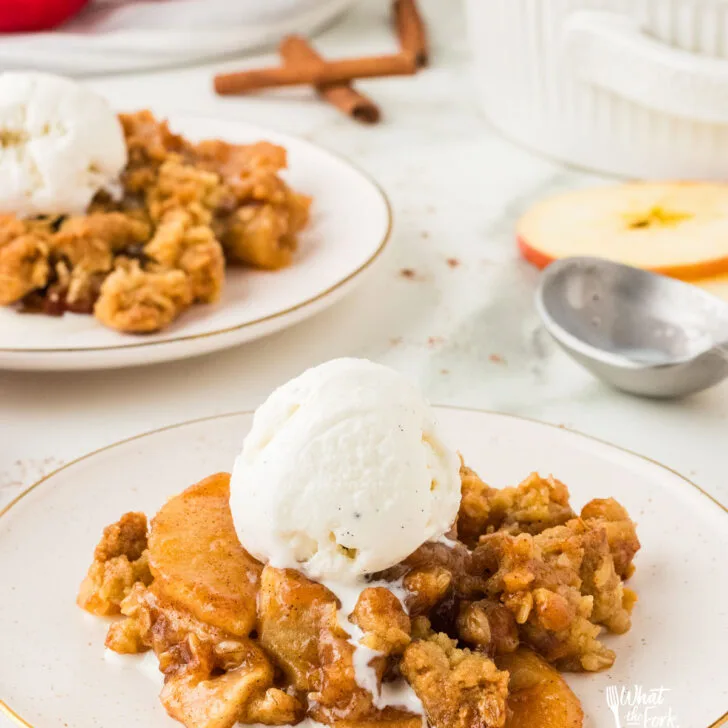 a serving of gluten free apple crisp on a round white plate topped with a scoop of vanilla ice cream. Another plate of the same is in the background
