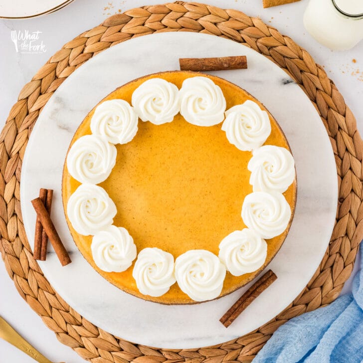 overhead shot of a Layered Pumpkin Cheesecake made with a Gluten Free Crust and decorated with dollops of whipped cream served on a white marble platter on a rattan charger.