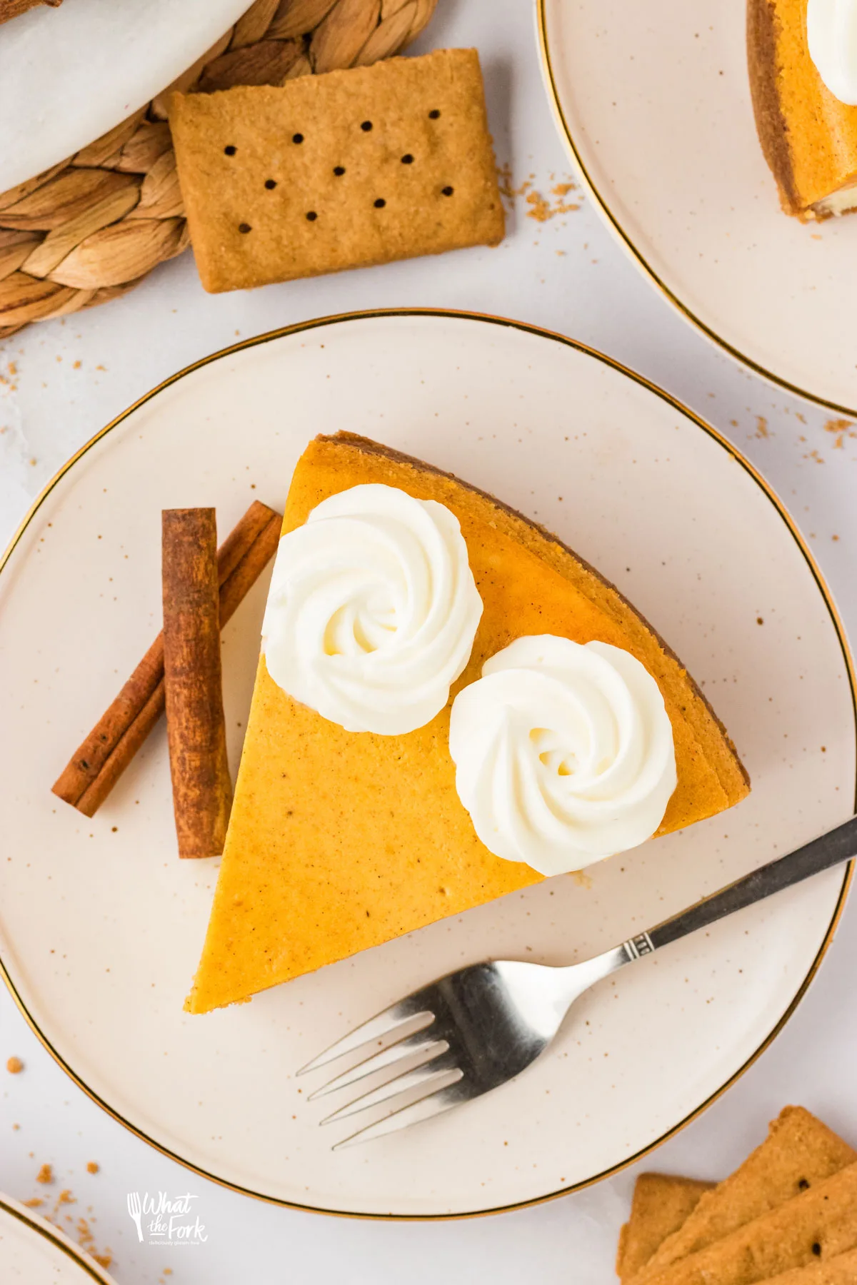 overhead shot of a slice of Layered Pumpkin Cheesecake with a Gluten Free Crust on a small round white plate with a silver fork and two cinnamon sticks on the side