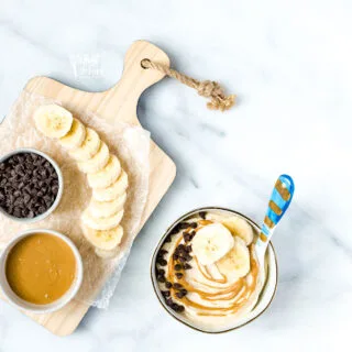 1-Ingredient Banana Nice Cream in a small bowl with a blue spoon topped with sliced bananas, drizzled peanut butter, and mini chocolate chips