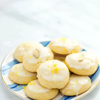 a plate of soft baked gluten free lemon ricotta cookies on a white and blue striped plate. The cookies are topped with lemon glaze and some of the cookies are decorated with lemon zest or a slice of almond.