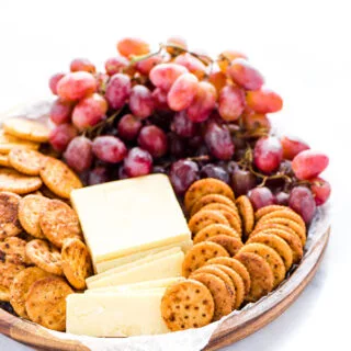 a wood platter lined with wax paper and topped with a block of cheddar, some cheddar slices, red grapes, and gluten free seasoned crackers