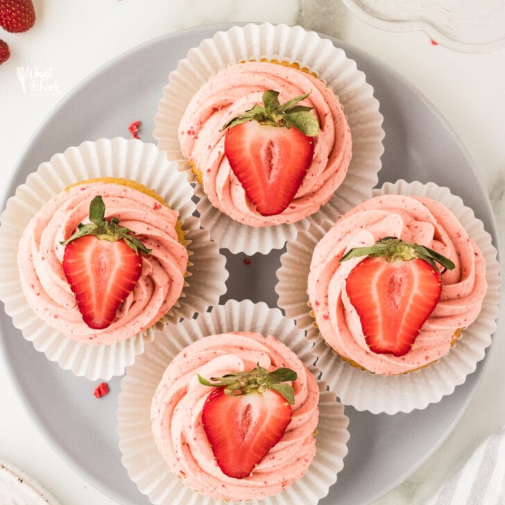 overhead shot of 4 cupcakes on a round white plate frosted with a swirl of strawberry frosting and topped with half of a strawberry
