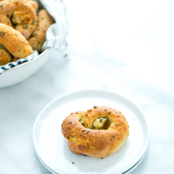 baked Gluten Free Garlic Knots in a serving bowl in the background with one garlick knot on a small round white plate in the front