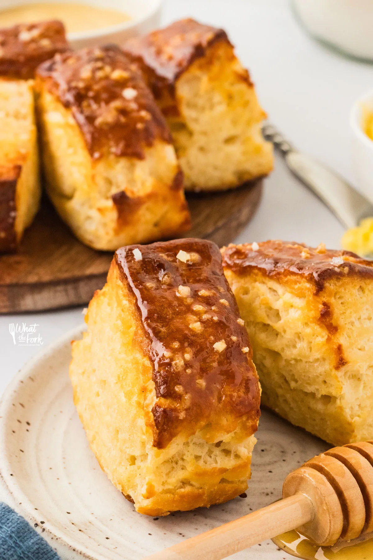 two Buttery Pretzel Biscuits topped with pretzel salt on top of a small white pottery plate with a honey spoon with more pretzel biscuits in the background