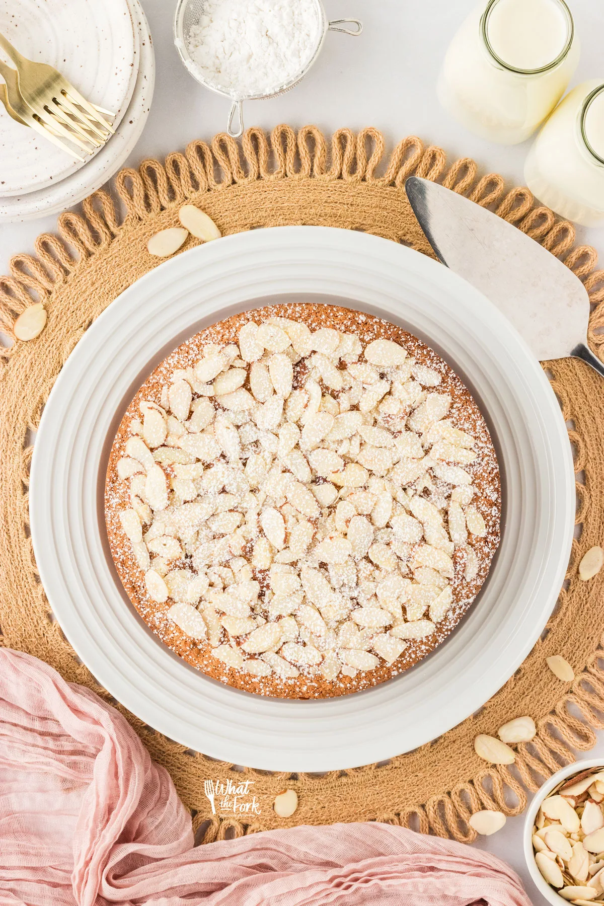 overhead shot of a baked almond flour cake topped with sliced almonds and dusted with powdered sugar. The cake is on a round white cake plate on top of a rattan charger.