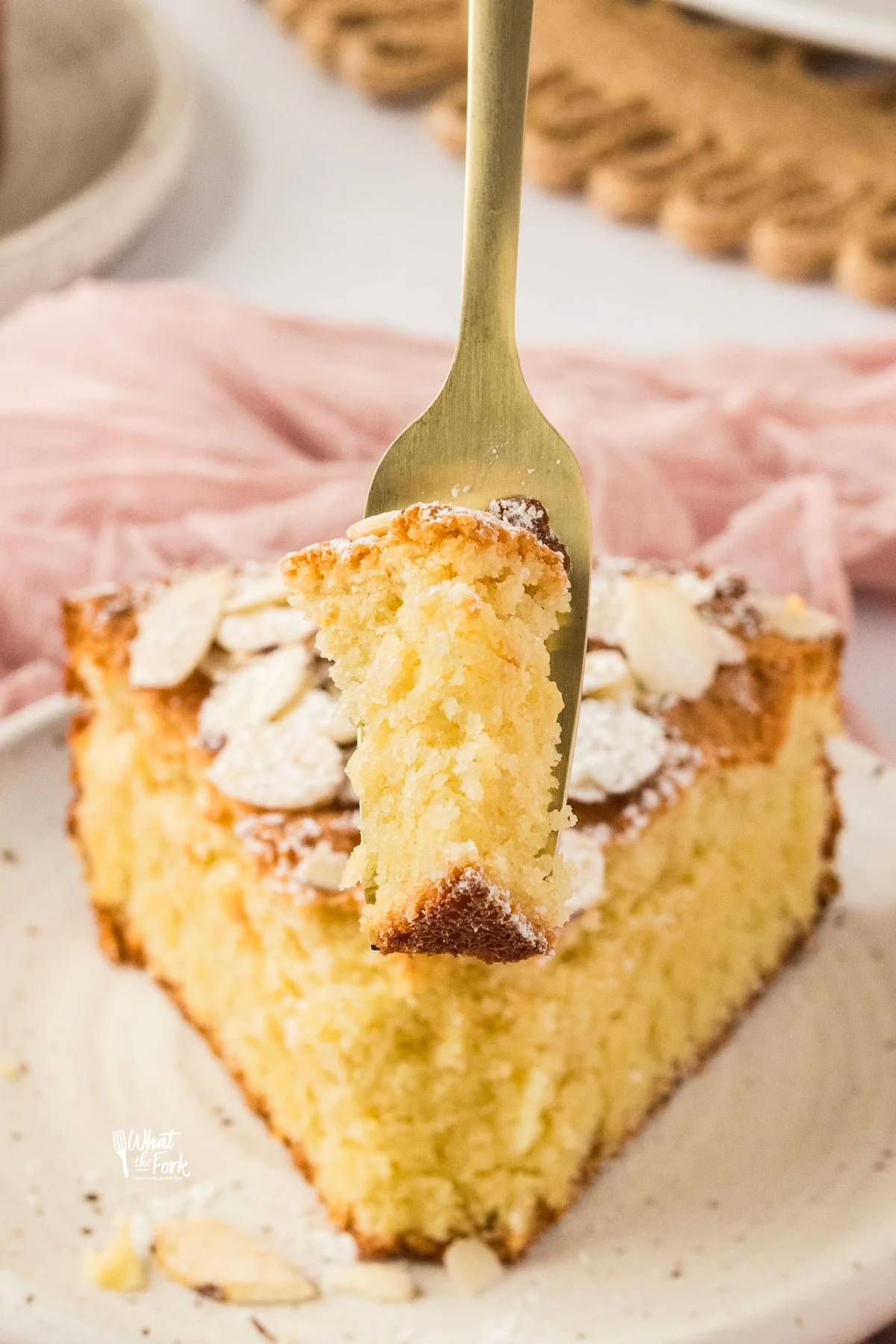 a slice of almond flour cake on a small round white plate with a forkful of cake held aloft with a gold fork for a bite