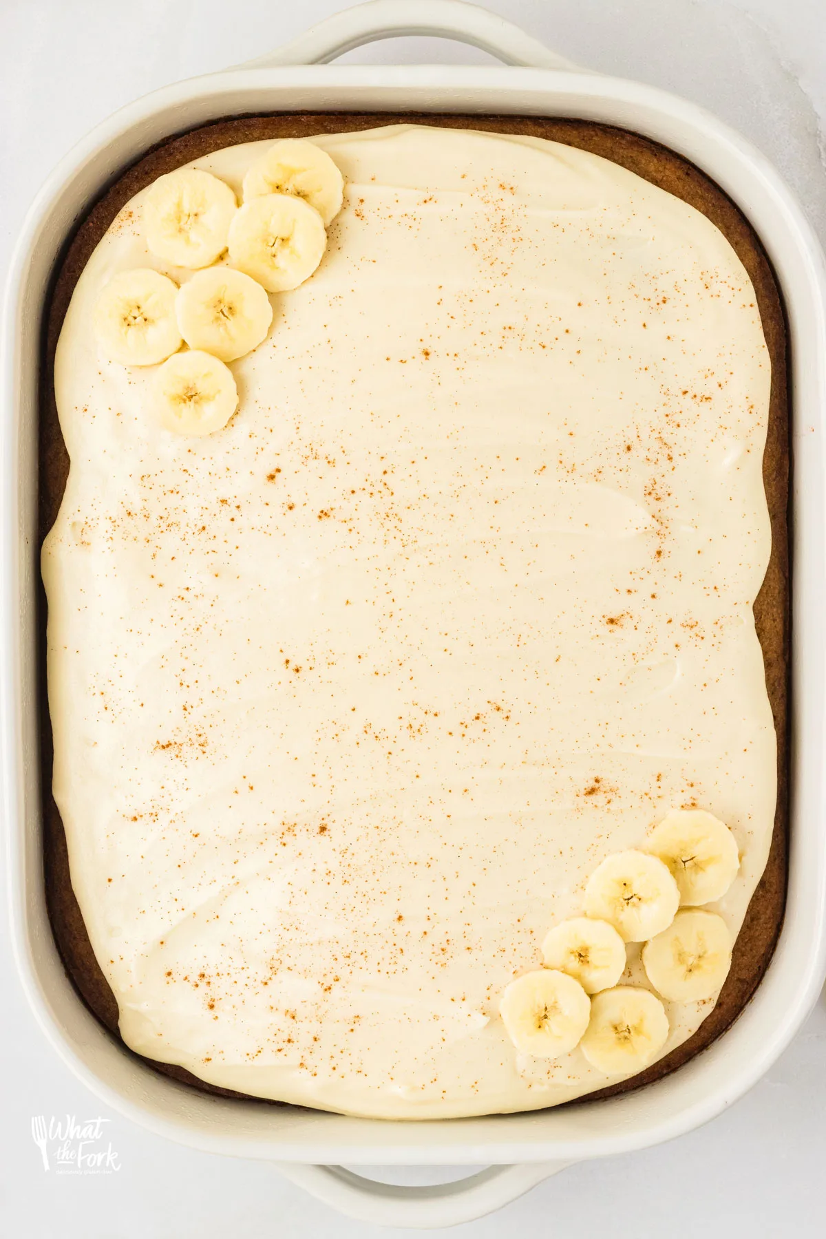 Gluten Free Banana Cake with Brown Butter in a 9x13 white baking pan frosted with maple cream cheese frosting and decorated with a dusting of cinnamon and banana slices