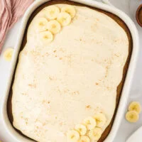 overhead shot of a Gluten Free Banana Cake with Brown Butter in a white 9x13 pan frosted with maple cream cheese frosting and garnished with a sprinkle of cinnamon and fresh banana slices.