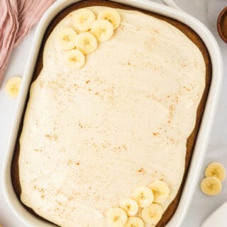 overhead shot of a Gluten Free Banana Cake with Brown Butter in a white 9x13 pan frosted with maple cream cheese frosting and garnished with a sprinkle of cinnamon and fresh banana slices.