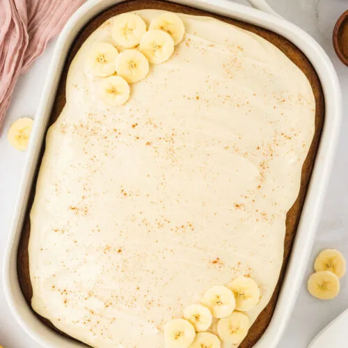 overhead shot of a Gluten Free Banana Cake with Brown Butter in a white 9x13 pan frosted with maple cream cheese frosting and garnished with a sprinkle of cinnamon and fresh banana slices.