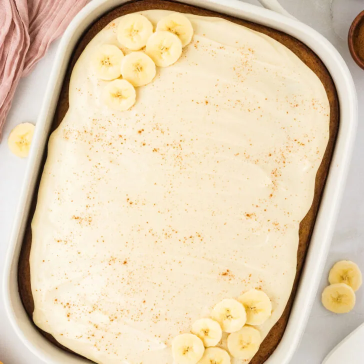 overhead shot of a Gluten Free Banana Cake with Brown Butter in a white 9x13 pan frosted with maple cream cheese frosting and garnished with a sprinkle of cinnamon and fresh banana slices.