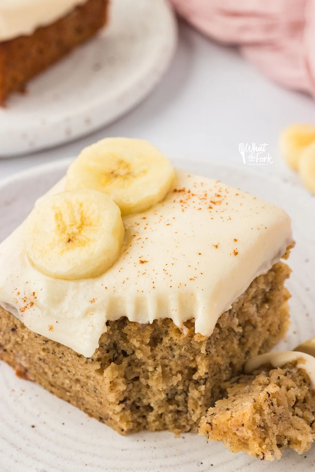 a slice of Gluten Free Banana Cake on a white plate topped with cream cheese frosting and banana slices with a forkful removed