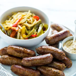 Slow Cooker Sausage and Peppers made with a secret ingredient. The sausages are cooked and plated on a white plate with a bowl of onions and peppers and a small bowl of stone ground mustard.