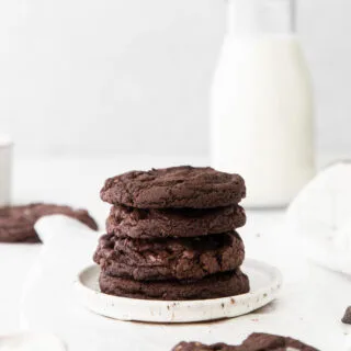 a stack of Gluten Free Chocolate Crunch Cookies on a small white plate with one in the foreground and a small glass of milk in the background