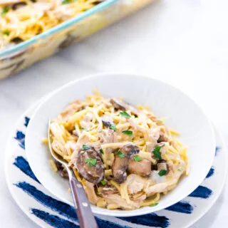 a small white plate of Gluten Free Chicken Tetrazzini with Rotisserie Chicken on top of a white and blue striped plate. The full casserole dish is behind.