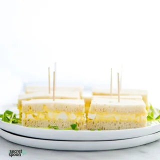 gluten free egg and mayo sandwiches with toothpicks on a stack of white plates garnished with baby cress